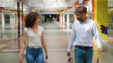 Çok ırklı genç bir çift alışveriş merkezlerini geziyor, alışveriş torbaları taşıyor, birlikte yürürken neşeli sohbetler yapıyor ve hafta sonları boş zamanların tadını çıkarıyor.