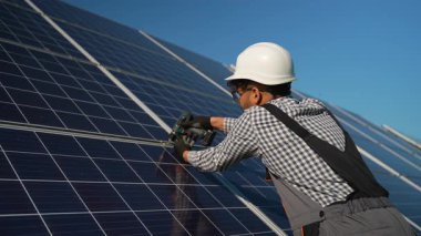 Güvenlik kaskı takan ve üniforma giyen profesyonel teknisyen açık mavi gökyüzü altında büyük ölçekli bir güneş çiftliğine fotovoltaik güneş panelleri kuruyor.
