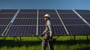 Güneş panelleri yakınında yürürken alet çantası taşıyan bakım mühendisi, parlak güneş ışığı altında fotovoltaik güç istasyonunda rutin kontrol yapıyor.