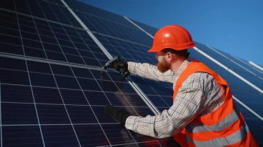 Turuncu kasklı ve yüksek görüşlü yeleği olan mühendis, açık mavi gökyüzünün altındaki büyük bir yapıya güneş panelleri kurmak için bir matkap kullanıyor.