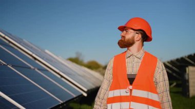 Geniş güneş çiftliğinde detaylı güneş paneli incelemesi yapan profesyonel mühendis parlak mavi gökyüzü altında sürdürülebilir enerji teknolojisi gösteriyor.