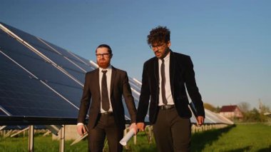 Güneş panelleri yakınında iş birliği yapan profesyonel giyimli işadamları, geniş kapsamlı yenilenebilir enerji alanı ayarlarında proje ayrıntılarını planlar ve dijital tabletle gözden geçiriyorlar