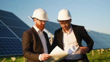 Şapkalı ve takım elbiseli iki mühendis bir güneş enerjisi santralinin planlarını inceliyor, açık gökyüzü altında planlar ve ilerlemeyi tartışıyorlar.