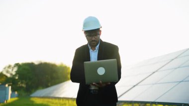 Güneş enerjisi uzmanı fotovoltaik panellerin yanında duran koruyucu ekipmanları giyiyor, yenilenebilir enerji santralinde teknik denetim yaparken dizüstü bilgisayardaki verileri analiz ediyor