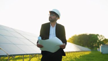 Güneş paneli planlarını inceleyen, güneşli alanda duran, yenilenebilir enerji projesinin gelişimini analiz eden, başlık takan profesyonel bir mühendis.