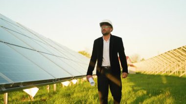 Beyaz kasklı ve siyah elbiseli Arap mühendis güneş panellerinin yanında güvenle yürüyor, geniş yenilenebilir enerji alanının teknik planlarını taşıyor.