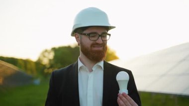 Profesyonel yenilenebilir enerji mühendisi geniş güneş paneli çiftliğinde dururken ampulü tutuyor, yenilikçi sürdürülebilirlik yayıyor ve izleyiciye güvenle gülümsüyor.