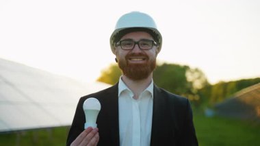 Güneş enerjisi santralinin yanında duran, gün batımında aydınlatılmış ampulü tutan, sürdürülebilir enerji yeniliğini temsil eden, sabit şapka ve iş kıyafeti giyen mühendis.