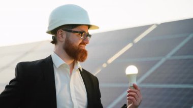 Kendine güvenen kızıl saçlı kadın mühendis güneş paneli kurulumunun yanında duruyor. Parlayan ampul, yenilikçi yenilenebilir enerji çözümlerini temsil ediyor.