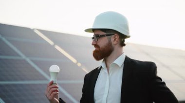 Yenilenebilir enerji mühendisi büyük bir güneş paneli çiftliğinin önünde ampul tutuyor, gülümsüyor ve kameraya bakıyor, farklı ifadeler gösteriyor.