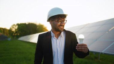 Kendine güvenen profesyonel mühendis yenilenebilir enerji buluşunu vurguluyor, güneş panellerinin yanında altın gün batımında ampulle duruyor.