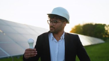 Gıcır gıcır iş kıyafeti giyen profesyonel mühendis yenilenebilir enerji tesisinde altın gün batımı ışığı altında güneş panellerinin yanında güven içinde duruyor.