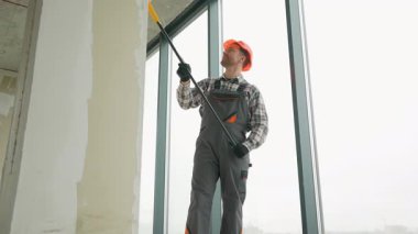Gri tulumlu ve turuncu kasklı inşaat işçisi inşaat halindeki yeni bir binanın büyük penceresinin yanına beyaz bir duvar boyuyor.