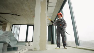 Profesyonel ressam titizlikle beton duvara astar takıyor, aktif inşaat alanında turuncu güvenlik kaskı ve gri üniforma giyerken uzun silindir kullanıyor.