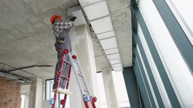 Modern bir ofis binasında tavan monte edilmiş havalandırma kanalı üzerinde çalışan işçi tulumu ve eldivenleri var..