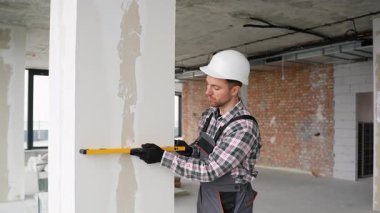 İnşaatçı, duvarın dikey düzlüğünü dikkatlice kontrol ediyor hassas apartman yenileme sırasında ruh seviyesini kullanıyor, profesyonel miğfer takıyor.