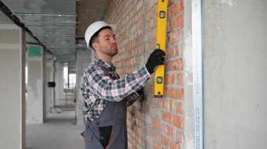 Güvenlik kaskı takan profesyonel müteahhit inşaat alanında çalışırken ruh seviyesini kullanarak tuğla duvarın dikeyliğini dikkatlice kontrol ediyor.