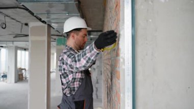 Profesyonel inşaat işçisi güvenlik donanımı takıyor tuğla duvar ve bitmemiş bina projesinin içinde mezurayla ölçüm yapıyor, mimari gelişimde hassasiyet ve beceri gösteriyor.