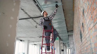 Üniformalı teknisyen, verimli hava akımı ve teknik hassasiyeti sağlamak için modern bir ofise dikkatlice tavan havalandırma ünitesi kuruyor..