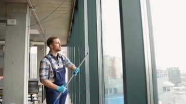 Mavi eldiven ve tulum giyen tamirci, çağdaş ofis alanındaki büyük cam pencereleri dikkatle temizliyor ve pırıl pırıl ve pırıl bir manzara sağlıyor.
