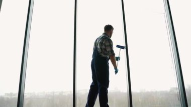 Profesyonel pencere temizleyicisi mavi tulum ve eldiven giyerek modern ofis binasında büyük panoramik pencereleri dikkatle yıkar ve tertemiz ve pırıl pırıl parlar.