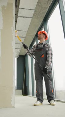 Gri üniformalı profesyonel bir ressam ve turuncu kasklı. Duvarlara ve boş ofis alanının tavanına astar takıyor. Geniş pencereli.