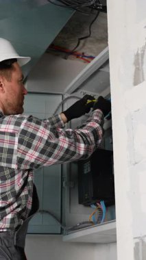 Güvenlik kaskı ve eldiven takan profesyonel elektrikçi elektrik paneli üzerinde çalışıyor, kabloları ve çeşitli elektrik bileşenlerini dikkatlice yerleştiriyor.