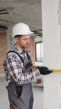 Koruyucu iş giyiminde yetenekli bir müteahhit, inşaat projesi sırasında duvarın ruh seviyesini dikkatlice kontrol ederek, tam olarak dikey konumlandırmayı sağlıyor.