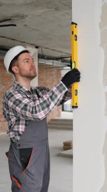 Üniforma ve şapka giyen profesyonel müteahhit, bina içinde yeni inşa edilen duvarın dikey hizalanmasını sağlamak için ruh seviyesini kullanır.