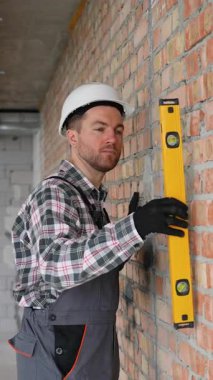 Üniforma ve şapka giyen profesyonel müteahhit bina inşaatı sırasında bir tuğla duvarın doğru yüksekliğini sağlamak için ruh seviyesini kullanır.