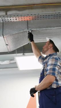 Koruyucu üniforma ve güvenlik kaskı takan inşaat işçisi HVC sistem bakımı sırasında profesyonel tornavida kullanarak metal hava kanalına yalıtım malzemesi yerleştiriyor.