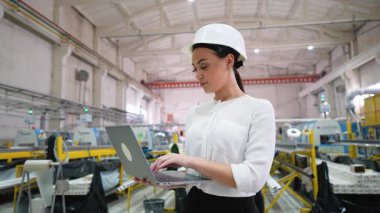 Genç bayan mühendis dizüstü bilgisayarda yazı yazan beyaz bir şapka takıyor, büyük bir endüstriyel fabrikadaki işlemleri yönetiyor ve üretim süreçlerini izliyor.