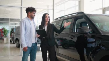 Profesyonel araba satıcısı, gösterişli siyah jip sunuyor. Potansiyel erkek alıcıyı, modern galeride araç özellikleri ve faydaları üzerinden yürütüyor.