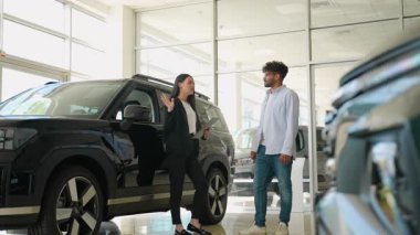 Modern bir araba galerisindeki bir erkek müşteriye yeni bir siyah arabanın özelliklerini sunan profesyonel satıcı, araç özellikleri ve satın alma seçenekleri hakkında tartışmalar yapıyor.