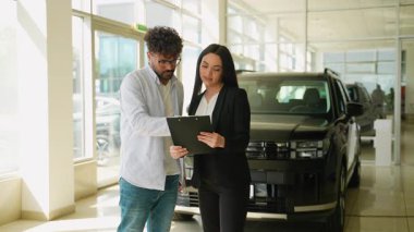 Profesyonel satıcı, bir araba galerisinde siyah bir cipin yanında duran bir müşteriye önemli belgeleri panoda gösteriyor.