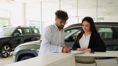Profesyonel araba satıcısı müşteriye son satın alma adımlarında rehberlik eder, evrak imzalar ve araba anahtarlarını modern galeride nakleder.
