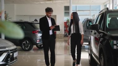 Profesyonel araba satıcısı potansiyel bir alıcıya çeşitli araçlar gösteriyor, özellikleri tartışıyor ve modern bir araba galerisinde bilgilendirilmiş bir karar vermesine yardımcı olacak bilgiler sağlıyor.