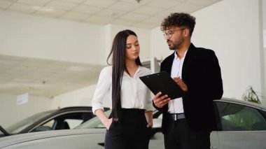 Profesyonel bir araba satıcısı, yeni bir araç almadan önce koşulları ve şartları tartışan bir kadın müşteriye galeride kontrat gösteriyor.