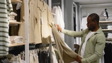 Modern perakende mağazasında şık erkek kıyafetleri seçerken, Afro-Amerikalı erkek müşteri giyim raflarına göz atıyor, gömlek ve pantolonları inceliyor.