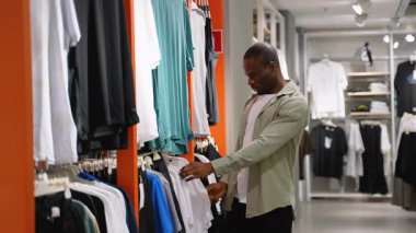 Kendine güvenen Afro-Amerikan erkek erkek giyim koleksiyonu, kumaş kalitesini ve tarzını dikkatlice değerlendirirken modern perakende mağazasındaki elbise raflarından gömlek seçiyor.