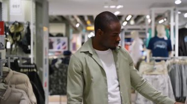 Genç Afro-Amerikalı erkek moda mağazasına giriyor, raflarda asılı gömlek ve pantolonları dikkatle inceliyor, kalite kontrolü yapıyor ve kıyafet seçerken fiyat etiketlerini karşılaştırıyor.