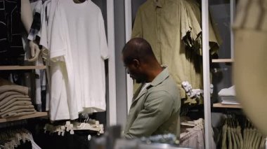 Kendine güvenen genç profesyonel beyaz t-shirt 'ü dikkatle inceliyor, kumaş kalitesini ve uyumu değerlendirirken moda perakende giyim mağazasının iç kısımlarına göz atıyor