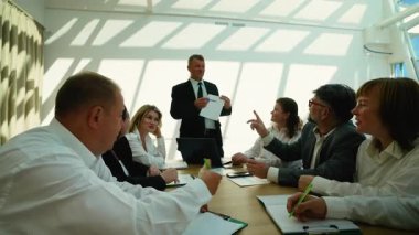 Üst düzey erkek patron, bir şirket brifingi sırasında meslektaşlarına yeni bir proje planı sunuyor. Özenli iş adamları modern güneşli bir konferans salonunda masada oturuyorlar.
