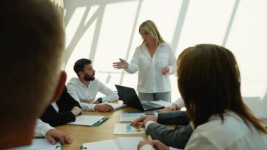 Modern bir ofiste ayakta duran ve bir iş toplantısına liderlik eden kendine güvenen kadın yönetici, konferans masasının etrafında oturan dikkatli meslektaşlarına yeni bir strateji sunuyor.