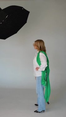 Profesyonel bir stüdyoda ışıklandırma ekipmanıyla bir fotoğrafçıya poz veren şık, orta yaşlı bir kadın beyaz arka planda bir moda markası için içerik yaratıyor.