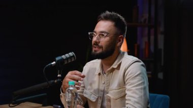 Beyaz tenli, sakallı ve gözlüklü bir radyo sunucusu profesyonel bir stüdyoda ses podcast 'i kaydederken duygusal bir hikaye anlatıyor. Canlı yayın sırasında elleriyle işaret ediyor.