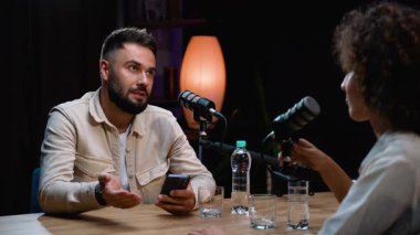 Profesyonel bir stüdyoda canlı yayın yapan iki kişi, kadın bir sunucunun akıllı telefonuna bakıp konuşma sırasında el kol hareketi yaparken sorularını cevaplayan bir erkek konuşmacıyla birlikte.