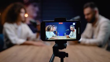 Bir tripoda akıllı telefon, canlı yayın ve sosyal medya içeriği oluşturulmasına odaklanmak için arka planı kasıtlı olarak bulanık olan bir erkek ve kadın konuk arasında bir podcast röportajı kaydediyor.