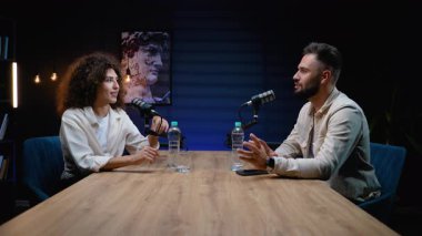 İki kişi, bir erkek ve bir kadın, birbirlerinin karşısındaki ahşap bir masada oturuyor ve bir podcast ya da radyo programı kaydediyor, profesyonel mikrofonları kullanıyor ve konuşurken işaret ediyorlar.