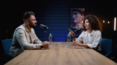 Profesyonel erkek ve kadın bir podcast kaydediyor veya modern bir stüdyoda canlı yayın yapıyor, mikrofonlu bir masada oturuyor ve meşgul edici bir röportaj sırasında aktif olarak bir konuyu tartışıyor.